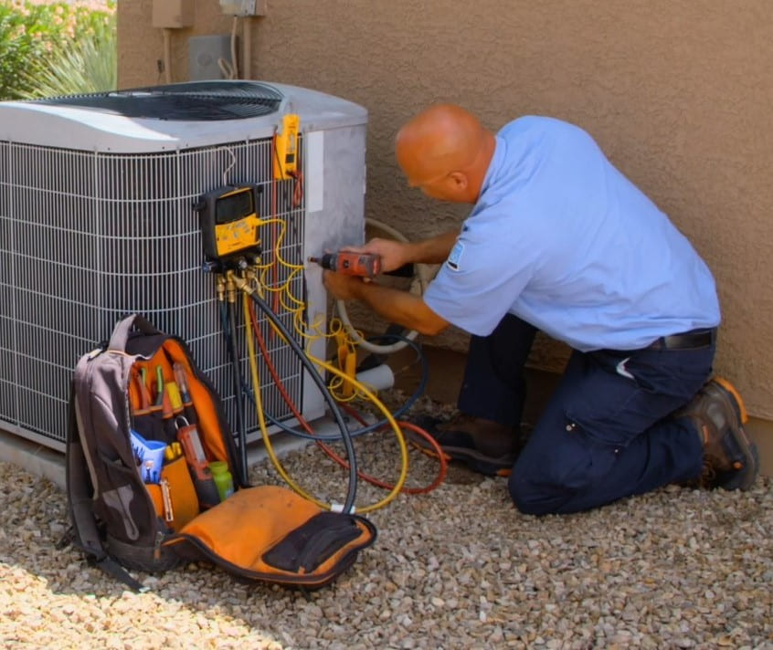 HVAC technician kneeling by outdoor air conditioner, using gauges and hoses to service the unit, with tool backpack nearby