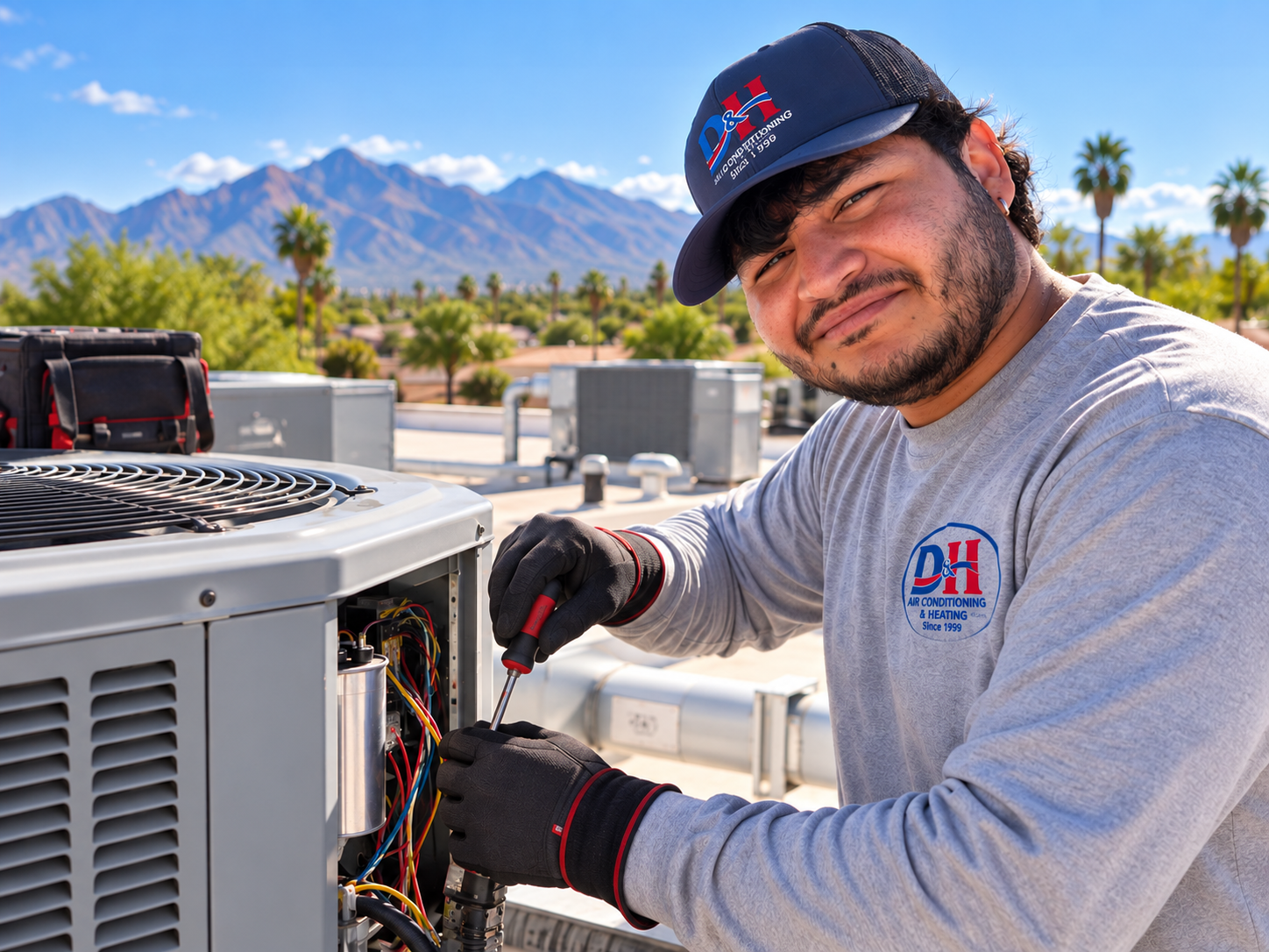 HVAC technician in a DH logo shirt repairs an outdoor air conditioner unit on a sunny rooftop, mountains in the background.