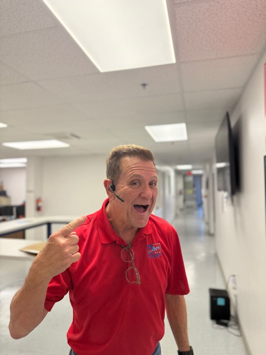 Smiling man in a red polo shirt with a headset, pointing to himself in a bright office hallway. Glasses hang from his shirt pocket.
