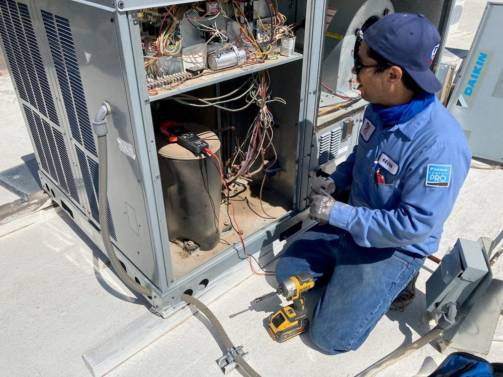 HVAC technician kneeling on a rooftop, servicing an open air conditioning unit with exposed wiring and a compressor inside, wearing a blue uniform and gloves.