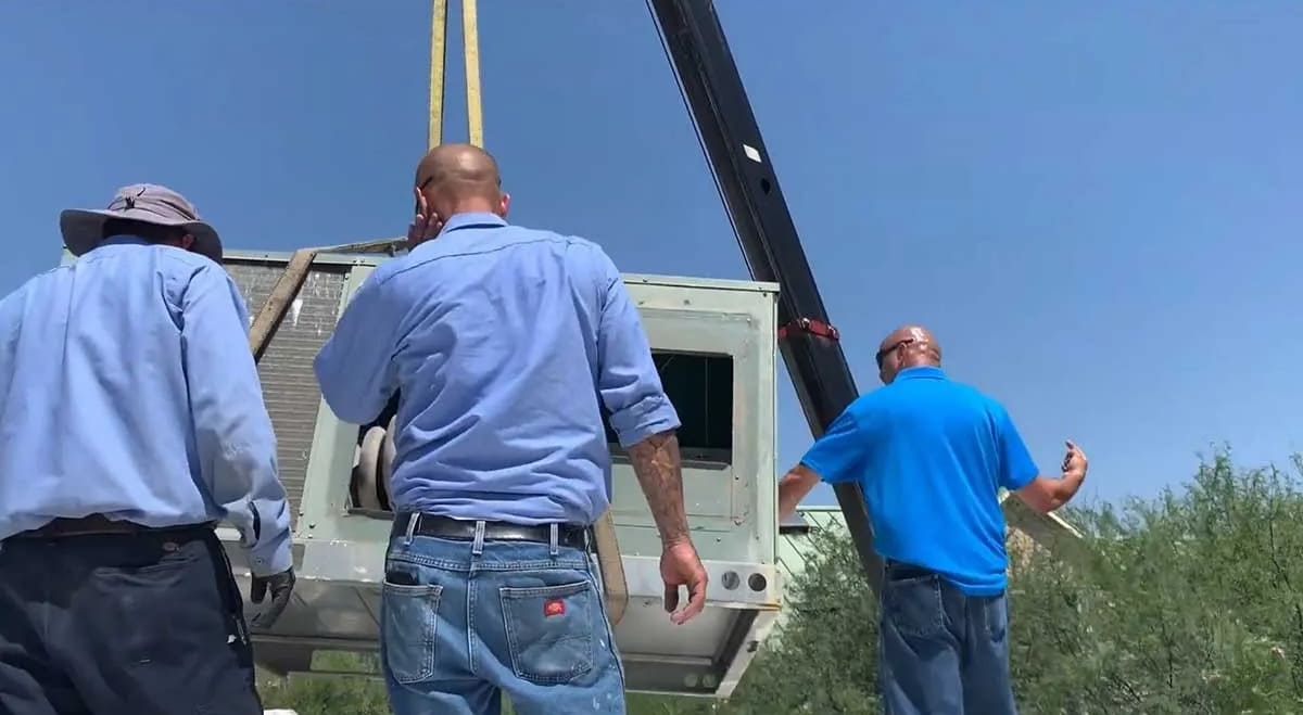 Three men in blue shirts and jeans guide a large HVAC unit being lowered by a crane outdoors on a sunny day.