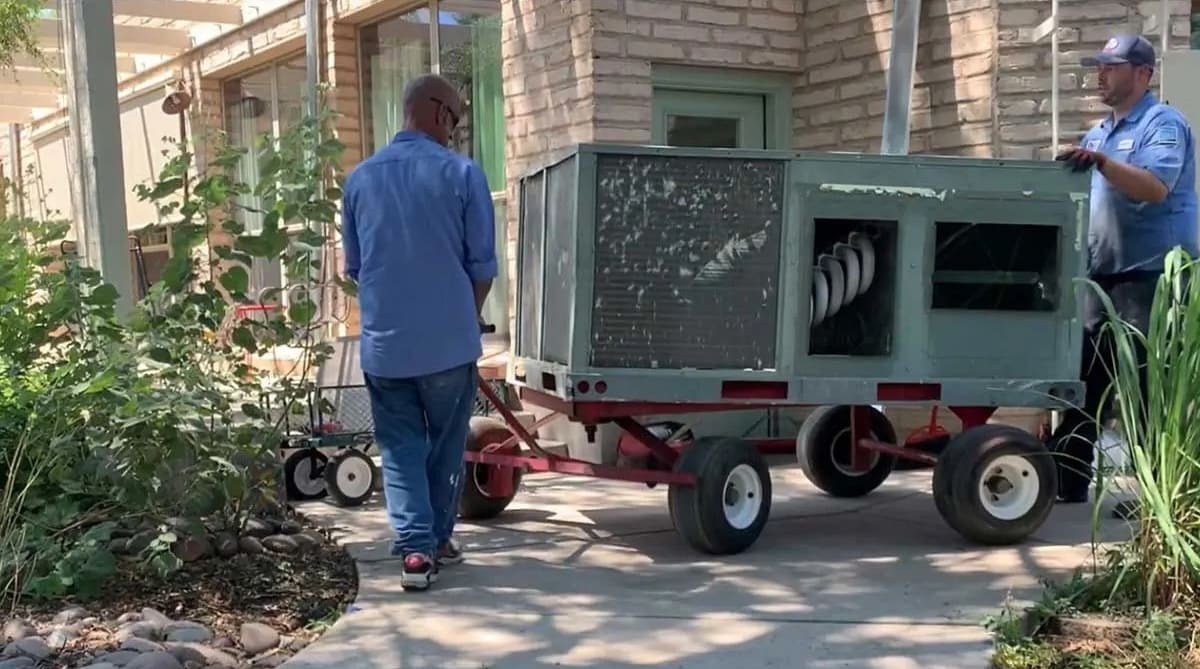 Two workers guide a large outdoor air conditioning unit on a trailer along a sidewalk with landscaping nearby, during a service call.