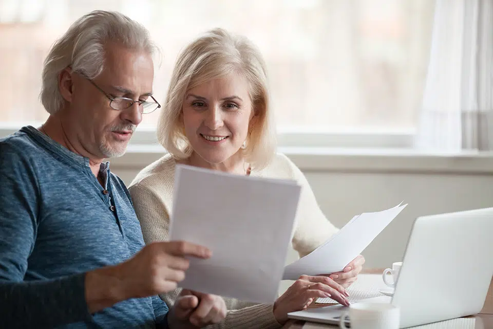 Older couple reviewing documents together at a bright table with a laptop nearby.
