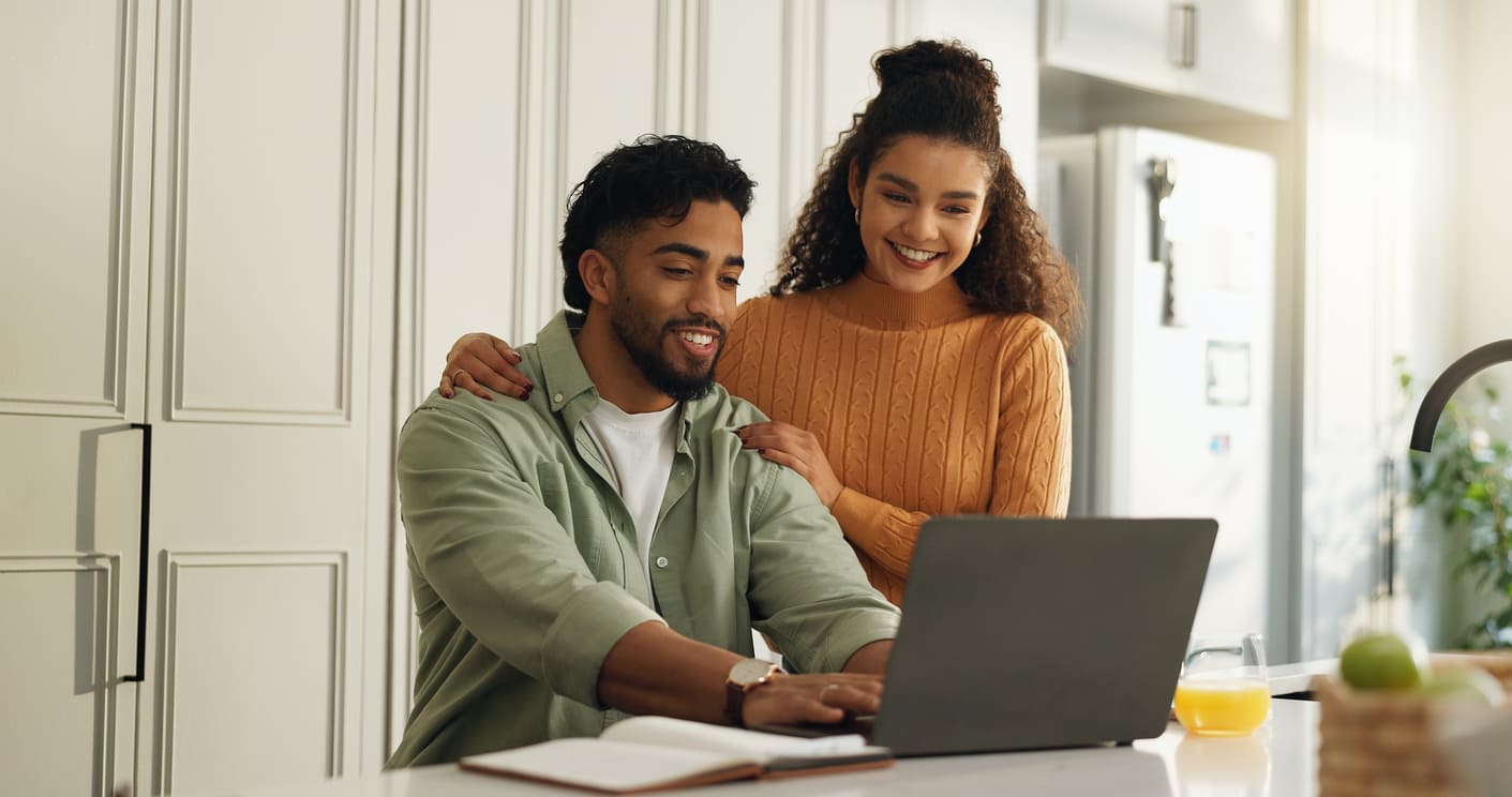 Couple in a bright kitchen, man typing on a laptop while woman stands beside him with a smile, resting a hand on his shoulder.