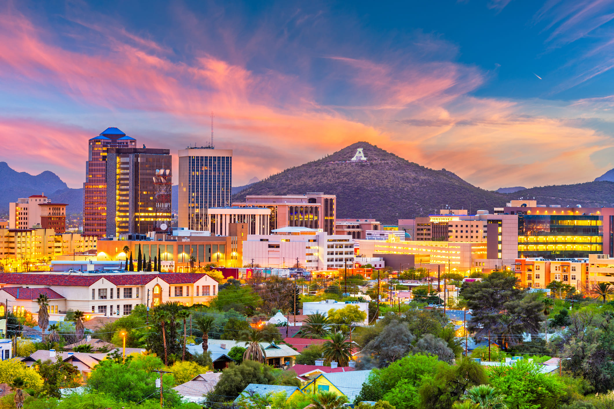 City skyline at sunset with pink-orange clouds, tall office buildings, and a hill bearing a white letter A.