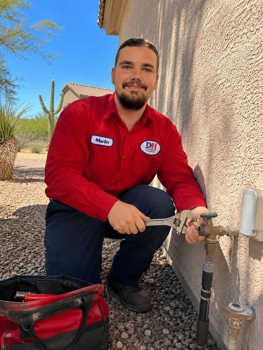 Technician in a red shirt kneels by a stucco wall, using a wrench on an outdoor faucet with a tool bag nearby