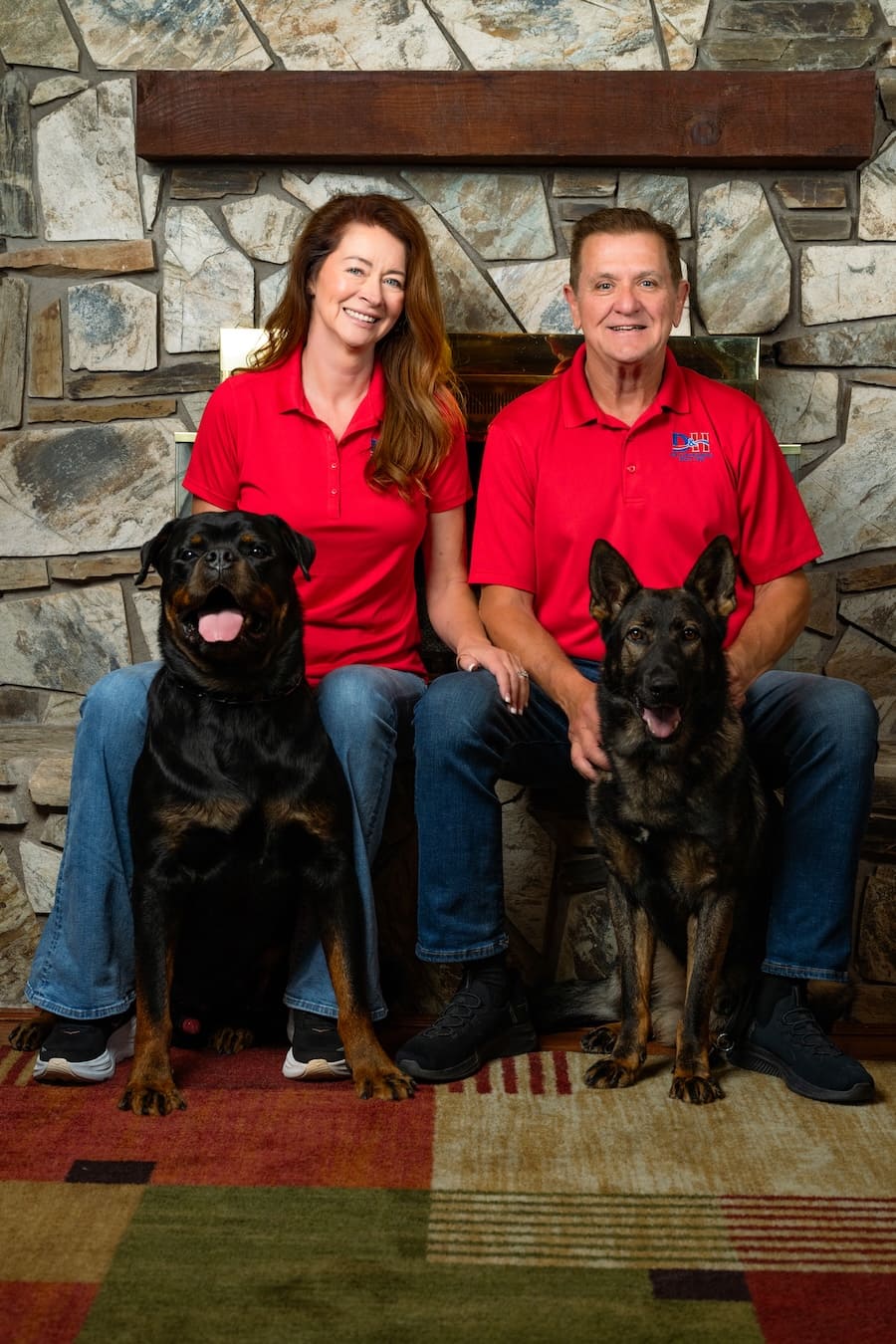 Couple in matching red polo shirts sit on a bench with two dogs in front of a stone fireplace, smiling at the camera.