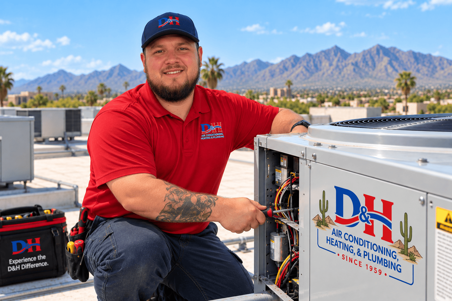 HVAC technician in a red polo and cap works on an outdoor air conditioner on a roof, mountains in the background, tool bag nearby, D&H branding visible on unit and shirt.